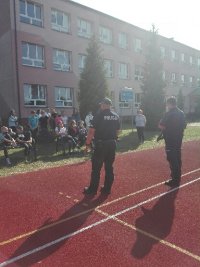 Spotkanie policjantów w szkole podstawowej w Poświętnem. Dzieci zebrane są na boisku szkolnym Policjanci stoją w odległości przed dziećmi. Rozmawiają o zagrożeniach