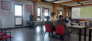posiedzenie Komisji bezpieczeństwa w Starostwie, policjantka z mikrofonem stoi przy stole, w tle widać prezentacje multimedialną oraz siedzące przy stołach osoby