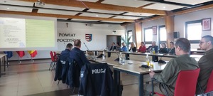 posiedzenie Komisji bezpieczeństwa w Starostwie, policjantka z mikrofonem stoi przy stole, w tle widać prezentacje multimedialną oraz siedzące przy stołach osoby