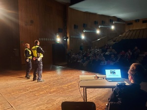 spotkanie z seniorami w  MDK w Opocznie na scenie stoją policjanci w tle widać projektor multimedialny,  przed sceną w fotelach siedzą osoby.