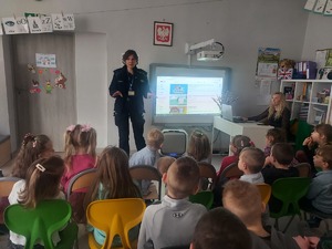 spotkanie z dziećmi. Policjantka stoi na środku klasy. W tle widać tablicę interaktywną. Dzieci siedzą na krzesełkach . Po prawej stronie przy biurku siedzi nauczycielka
