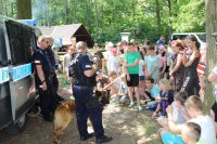 radiowoz policyjny oraz policjanci z psami sluzbowymi, policjant stoi przy radiowozie  trzyma psa sluzbowego na smuczy pies stoi  drugi policjant  stoi obok jego pies sluzbowy waruje. dzieci sluchaja zgromadzone obok