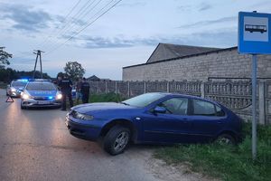 Asfaltowa droga, niebieski samochód, w tle policyjny radiowóz i policjanci pracujący na miejscu zdarzenia.