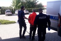 Umundurowani policjanci, prowadzą  zatrzymanego  za kradzież z włamaniem. Policjanci stoją tyłem  w środku stoi  zatrzymany, ręce na  skute kajdankami z tyłu. Z prawej strony widać policyjny radiowóz. w tle parking wewnętrzny.