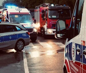Zdjęcie przedstawia, radiowozy wszystkich służb-policję, straż oraz pogotowie. na pierwszym planie po prawej stronie widać bok karetki pogotowia, w tle widać po lewej stronie  radiowóz policji, karetkę oraz straż pożarną.
