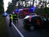 Zdjęcie wypadku drogowego w Anielinie. Na pierwszym planie widać dwa poważnie rozbite  auta w kolorze czarnym . Opel corsa  z rozbitym przodem oraz kia sportage z rozbitym przodem, powybijanymi szybami. W tle widać  pracujących na miejscu policjantów. Radiowozy straży pożarnej  oraz policji. obok las.