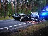 Zdjęcie wypadku drogowego w Anielinie. Na pierwszym planie widać dwa poważnie rozbite  auta w kolorze czarnym . Opel corsa  z rozbitym przodem oraz kia sportage z rozbitym przodem, powybijanymi szybami. W tle widać  pracujących na miejscu policjantów. Radiowozy straży pożarnej  oraz policji. obok las.