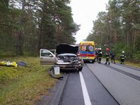 widać w tle stojący rozbity samochód w kolorze jasnym  samochód ma rozbity cały przód, stoi na pasie drogi, obok widać stojącego strażaka  w tle  radiowozy straży pożarnej