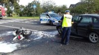 zdjęcie poglądowe z archiwum KPP wypadek drogowy z udziałem motocyklisty, motocykl leży na środku jezdni, obok stoi policjant  po prawej stronie na drodze stoi rozbity samochód osobowy w tle oznakowany radiowóz policji