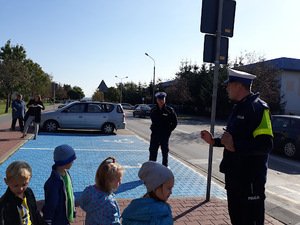 Ulica przy szkole. Po lewej stronie stoi policjant ruchu drogowego, rozmawia z uczniami klasy 1. Dzieci stoją przed policjantem po prawej stronie . W tle widać policjanta  oraz jezdnię i zaparkowany oznakowany radiowóz.
