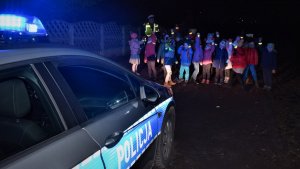 dzieci  na przejściu dla pieszych w kamizelkach, radiowóz oznakowany w tle dzieci w kamizelkach  i policjant