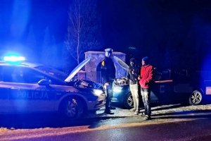 policjant  w mundurze stoi na przeciwko dwóch osob  obok zaparkowane pojazdy po lewej stronie radiowóz z otwarta maska w tle skoda z otwartą maską