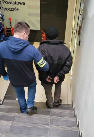 Policjanci prowadzą trzymając pod rękę zatrzymanych sprawców kradzieży . Zatrzymani mają na ręce trzymane z tyłu założone kajdanki