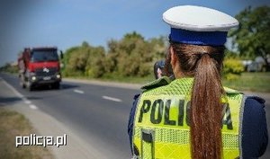 Na pierwszym planie znajduje się policjantka ruchu drogowego wykonująca pomiar prędkości.