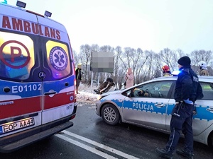 droga, stoją radiowóz i karetka. w polu w rowie leży na dachu rozbity samochód osobowy, Koło radiowozu stoi policjant. w tle strażacy