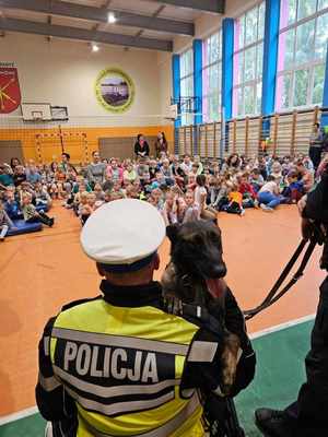 spotkania w szkołach. Policjanci stoją przed dziećmi, pokazują im radiowóz i motocykl służbowy. Na spotkaniu jest obecny przewodnik z spem służbowym.