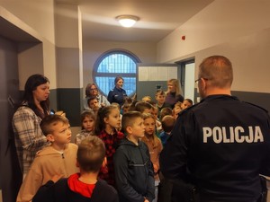 grupa dzieci i nauczycielka słuchają policjanta, policjantka stoi wśród dzieci w pomieszczeniu służbowym.