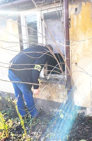 mężczyzna zagląda do domu przez wybite okno, na rękawie napis policja.