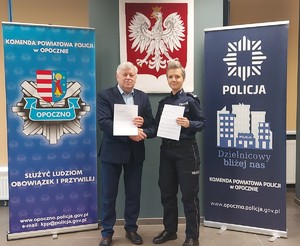 policjantka i mężczyzn w garniturze trzymają dokumenty i podają sobie ręce, za nimi godło, banery z napisem policja.