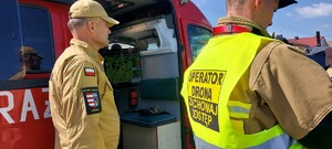 dwóch strażaków, fragment samochodu strażackiego, ekran, napis na kamizelce operator drona zachowaj odstęp.
