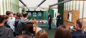 strzelnica, dwóch policjantów rozmawia z młodzieżą, prezentują broń.
