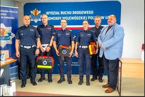 pięciu policjantów i mężczyzna stoją na tle napisu wydział ruchu drogowego, policjanci trzymają w rękach apteczki.