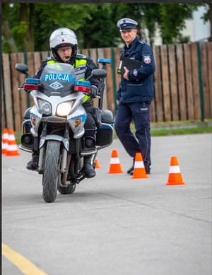 policjant jedzie motocyklem służbowym, za nim idzie policjant, na drodze rozstawione są pachołki.