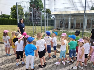 grupa dzieci stoi z policjantką przy ogrodzeniu.