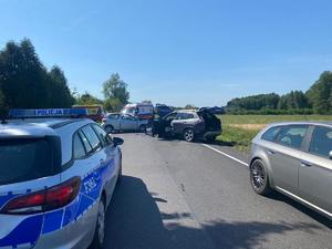 radiowóz policyjny, dwa rozbite samochody, w tle służby ratunkowe.