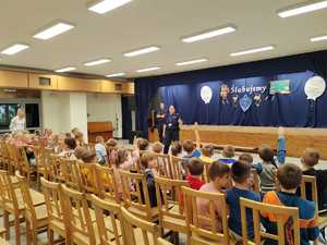 dzieci siedzą na krzesłach, policjanci stoją przed nimi, z boku stoi kobieta.