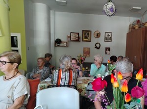 osoby starsze siedzą przy stołach, na ścianie wiszą ozdoby.