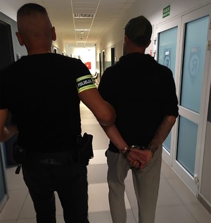 mężczyzna w opasce z napisem policja prowadzi mężczyznę w kajdankach.