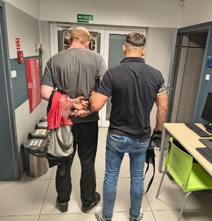 mężczyzna w kajdankach jest prowadzony przez mężczyznę z opaska z napisem policja.