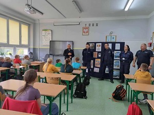 policjanci stoją w klasie lekcyjnej, uczniowie siedzą w ławkach.