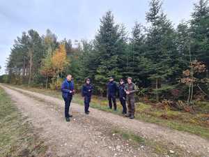 policjanci w mundurach, straż leśna w mundurach, mężczyzna w niebieskiej kurtce i z mikrofonem, wokół nich las.