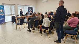 mężczyzna stoi, grupa ludzi siedzi na krzesłach, policjantka stoi za stołem.