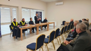 mężczyzna stoi za stołem, obok siedzą policjanci, przed nim grupa ludzi na krzesłach.