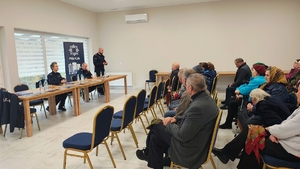 policjant w mundurze stoi przed grupą ludzi, którzy siedzą na sali na krzesłach, obok policjanci.