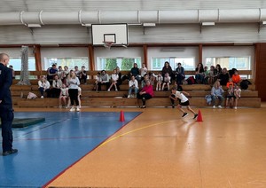 dzieci biegną na sali gimnastycznej, część dzieci siedzi na trybunach.