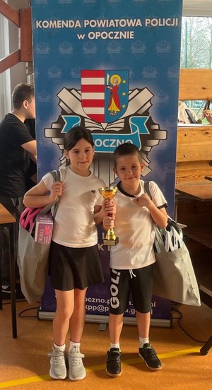 dwoje dzieci z pucharem i torbami stoją przed napisem komenda powiatowa policji w Opocznie.
