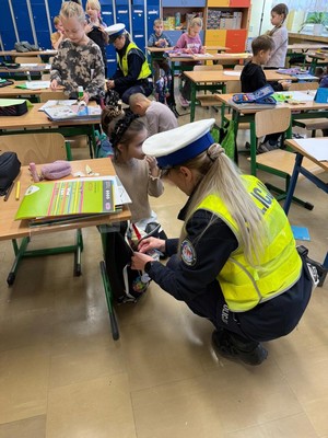 klasa lekcyjna, dzieci, policjantka zawiesza element odblaskowy do plecaka.