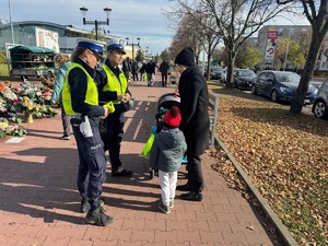 policjantki rozmawiają z kobietą z dzieckiem, za nimi kwiaty, chodnik.