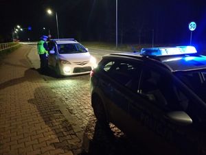 radiowóz, policjant stoi przy białym samochodzie, jedno światło w samochodzie się nie świeci.