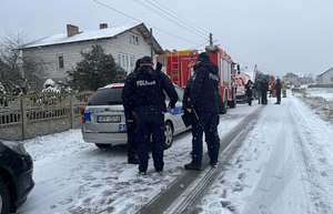 policjanci stoją na ośnieżonej drodze, samochody policyjne i strażackie, budynki.