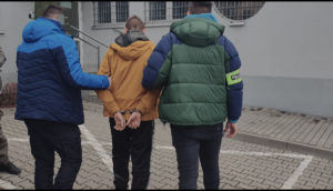 dwóch mężczyzn, jeden z opaską z napisem policja prowadzą mężczyznę w kajdankach na rękach trzymanych z tyłu.