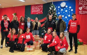 kobiet i mężczyźni, dzieci, część osób w czerwonych koszulkach, za nimi choinka.