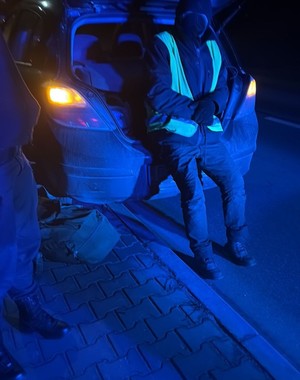 mężczyzna w odblaskowej kamizelce siedzi w otwartym bagażniku, ubrany w ciemne ubranie, obok widać stojącą osobę.