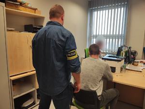 mężczyzna w niebieskiej koszuli stoi, dwóch męzczyzn siedzi przy biurku, jeden ma kajdanki na rękach.