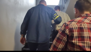 mężczyzna z opaską na ręce z napisem policja idzie z dwoma mężczyznami po schodach.