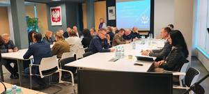 grupa ludzi, wśród nich policjanci i inne służby mundurowe siedzą przy stołach w sali konferencyjnej, na ścianie duży ekran.